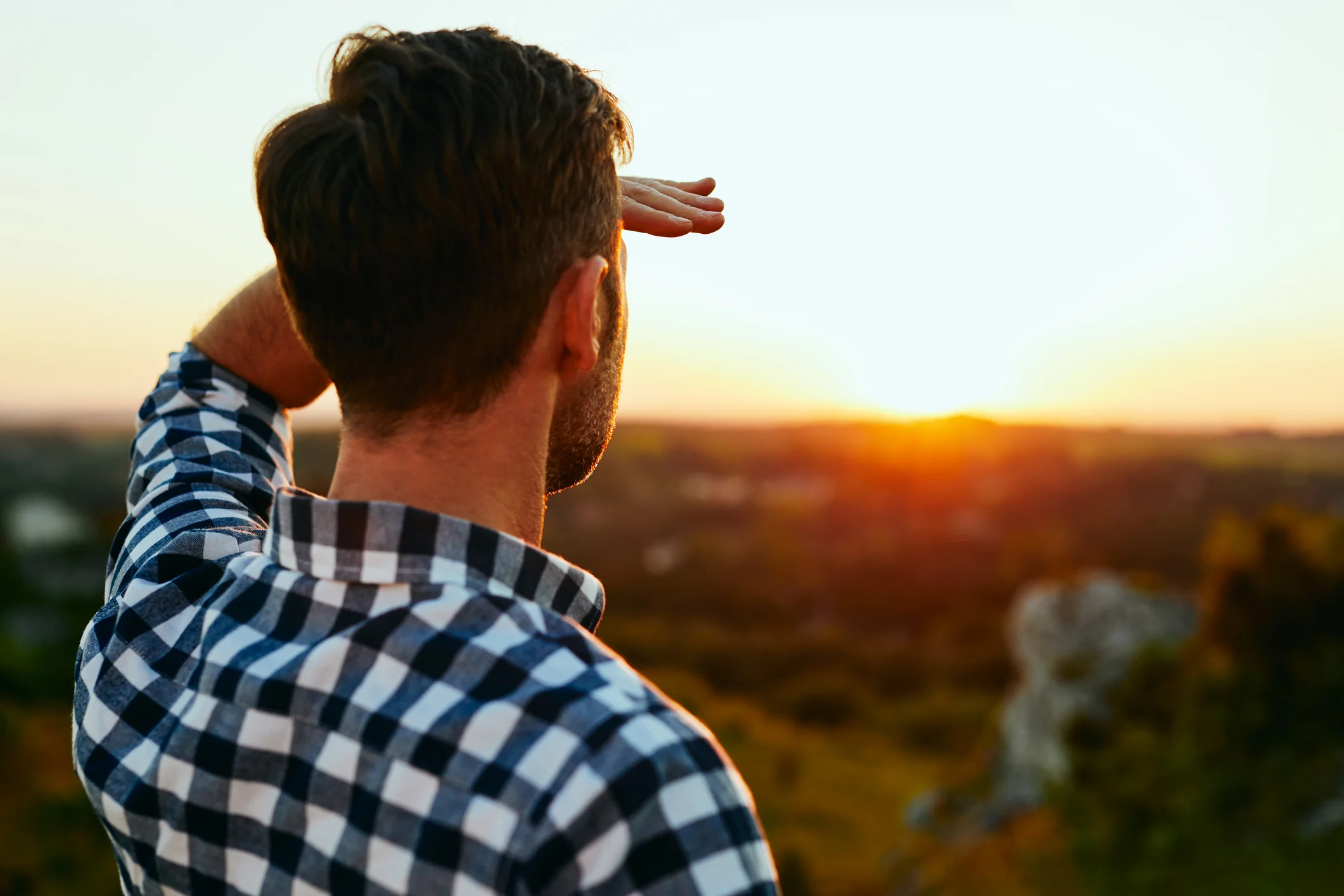 A man with his hand on his forehead looking out at a sunset over a landscape